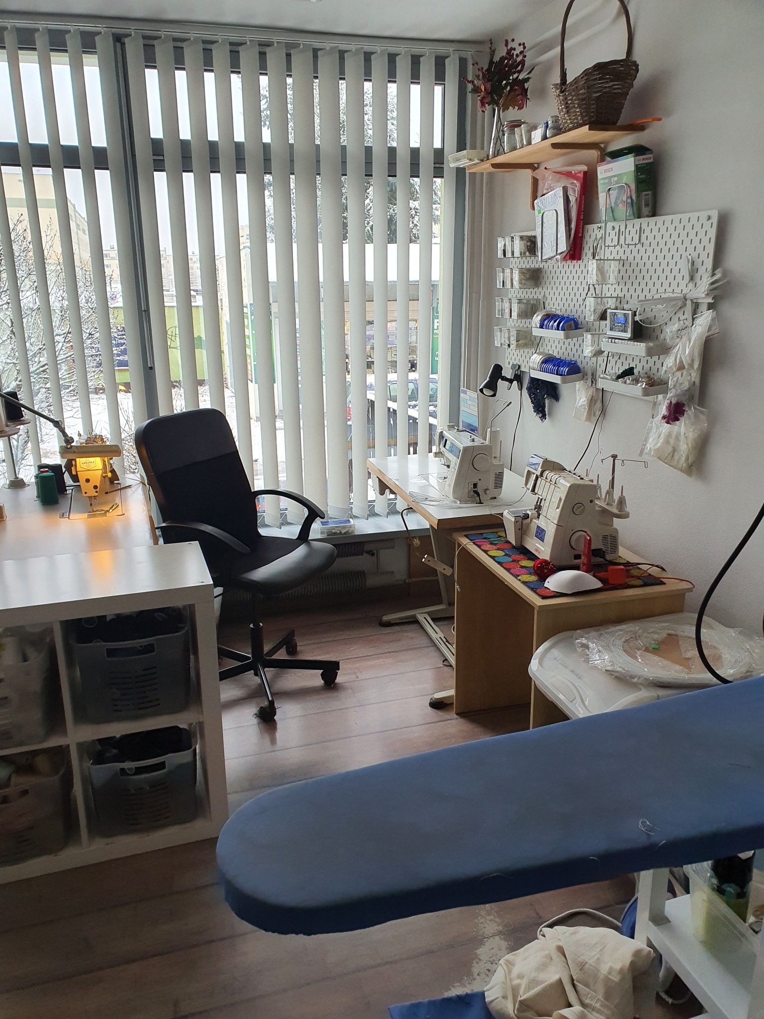 Pracownia krawiecka z dwoma maszynami do szycia, deską do prasowania i oknem z pionowymi żaluzjami.