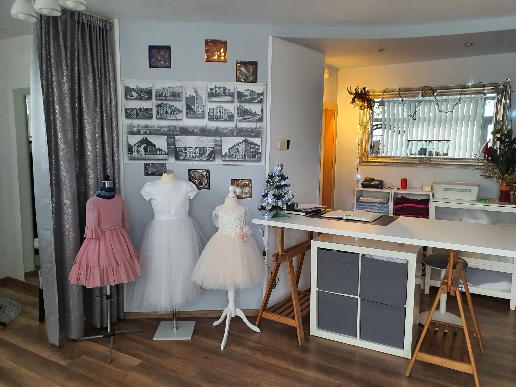 Pracownia krawiecka z trzema sukienkami na manekinach: różowa, biała i beżowa z koronką, na tle ściany z grafikami, biurka i dekoracji.