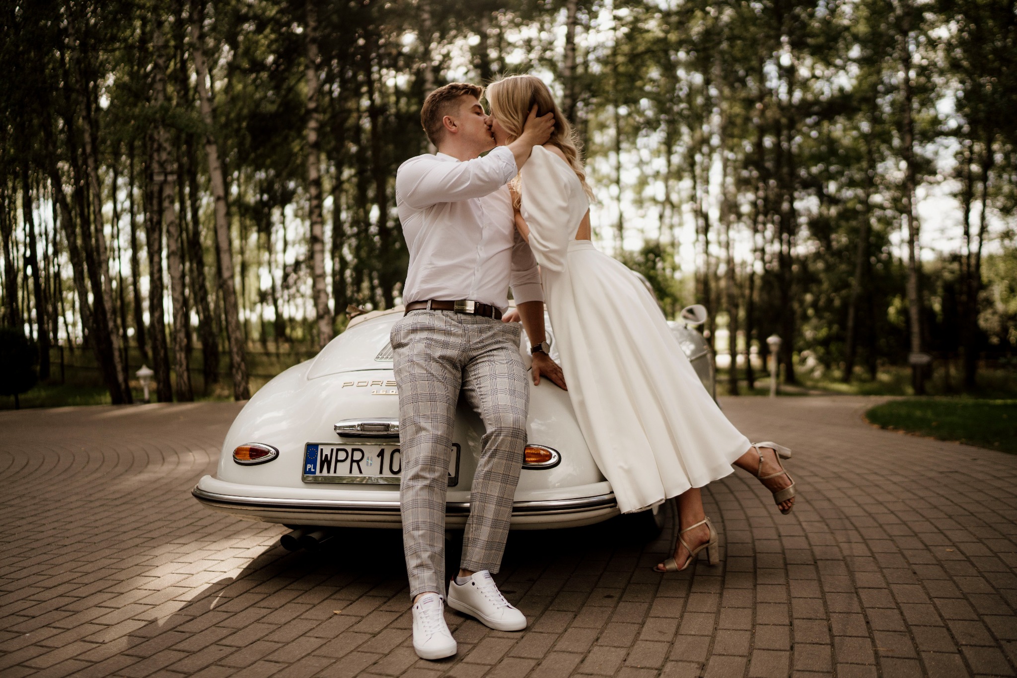 Para młoda całuje się opierając o białe Porsche 356 Speedster na tle drzew, panna młoda w sukni z odkrytymi plecami, pan młody w spodniach w kratę i białych sneakersach.