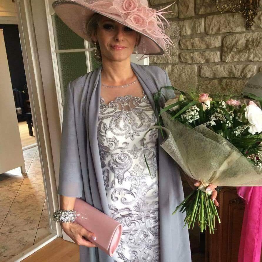 Kobieta w eleganckiej sukience z szarym kardiganem i kapeluszem z różami, trzymająca bukiet kwiatów i różową kopertówkę, pozuje na tle kamiennej ściany.