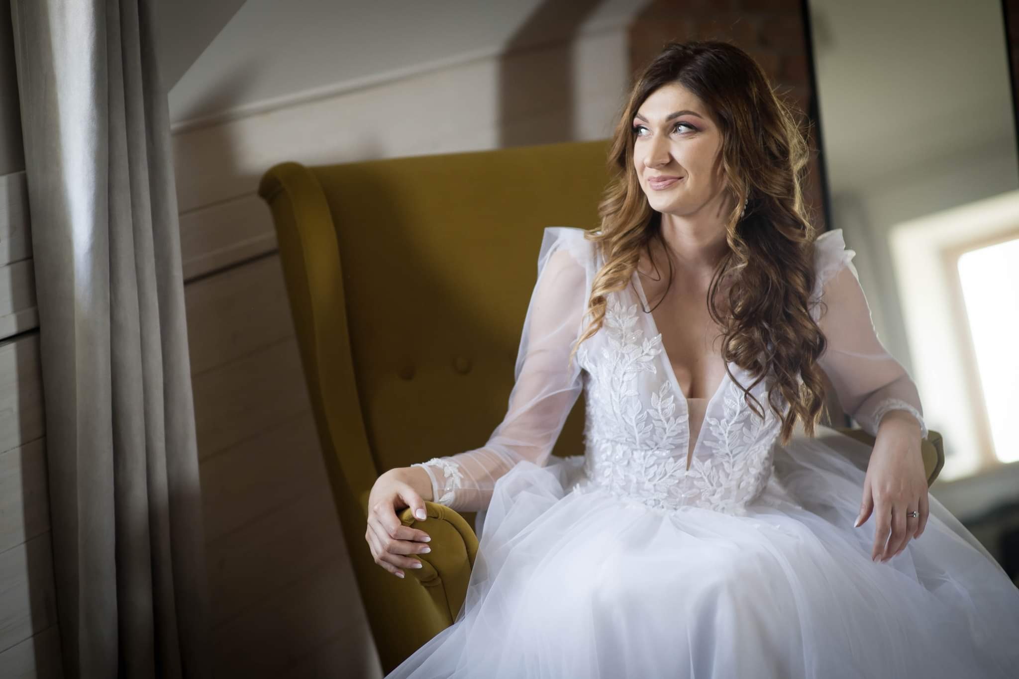 Panna młoda w białej sukni ślubnej z koronkowymi aplikacjami, siedząca w musztardowym fotelu w Pabianicach, spogląda w bok z uśmiechem.