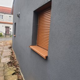 Polskiedachy - Elewacja budynku z szarą, ziarnistą strukturą, brązowe rolety zewnętrzne na oknach, widoczny fragment chodnika z popękanymi płytami.