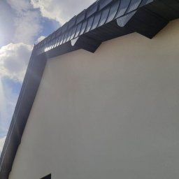 Polskiedachy - Fragment elewacji budynku z widocznym dachem pokrytym ciemną dachówką w kształcie łuski, oświetlonym jasnym słońcem i błękitnym niebem z chmurami w tle.