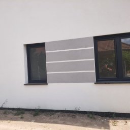 Polskiedachy - Elewacja budynku z dwoma czarnymi oknami, szarym pasem z białymi liniami i odbiciem fotografa w szybie.