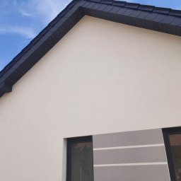 Polskiedachy - Fragment elewacji domu jednorodzinnego z widocznym dachem krytym ciemną dachówką, jasną elewacją i dekoracyjnym pasem szarości z białymi paskami pod oknem.