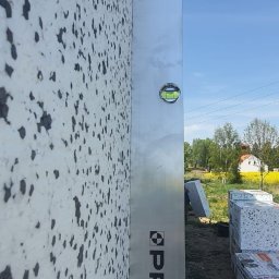 Polskiedachy - Ściana budynku pokryta styropianem, sprawdzana poziomicą marki PRO, z widocznymi paczkami styropianu w tle i fragmentem krajobrazu z domem.