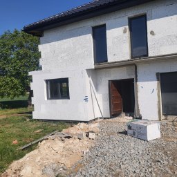 Polskiedachy - Surowy stan elewacji domu jednorodzinnego z białych bloczków, widoczne okna w ciemnych ramach, brama garażowa i częściowo wykonany dach, otoczenie placu budowy z gruzem i zielenią.