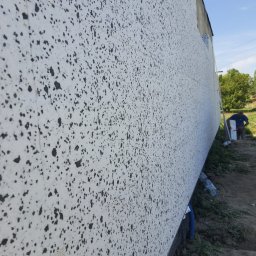 Polskiedachy - Ściana budynku w trakcie ocieplania, widoczne płyty styropianowe z nałożoną warstwą kleju z czarnymi plamkami, pracownik w oddali przycina płytę izolacyjną.