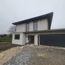 Polskiedachy - Surowy stan domu jednorodzinnego w trakcie budowy, widoczne białe bloczki, czarne okna i brama garażowa, na pierwszym planie gruz i ziemia, pochmurne niebo.