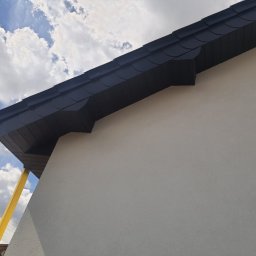 Polskiedachy - Fragment domu z nowym, ciemnym dachem z dachówki bitumicznej, widok z dołu na tle błękitnego nieba z chmurami. Żółta rura spustowa biegnie wzdłuż ściany.