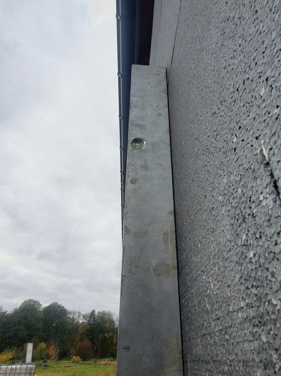 Szara ściana budynku w trakcie ocieplania, widoczny styropian i poziomica przy krawędzi, pochmurne niebo w tle.