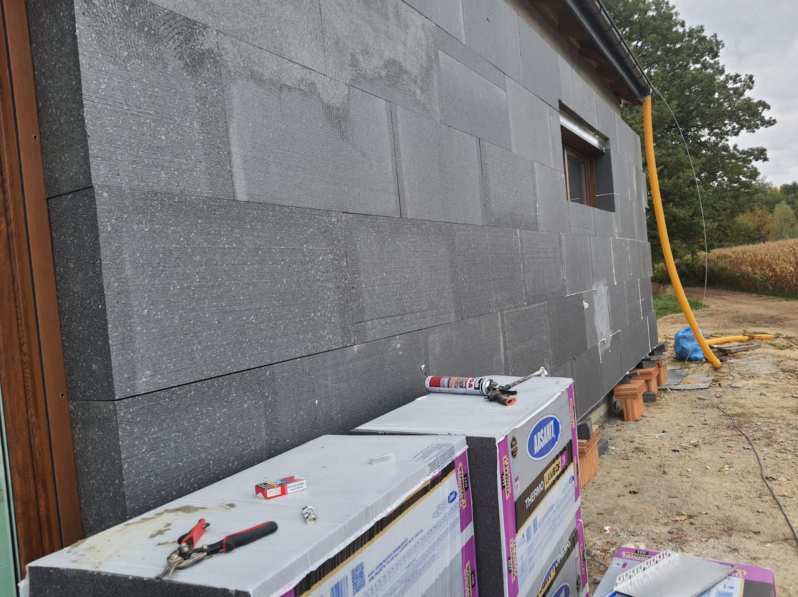 Ściana budynku w trakcie ocieplania, widoczne płyty termoizolacyjne z szarego styropianu, narzędzia pracy i materiały budowlane na pierwszym planie, żółta rura spustowa przymocowana do rynny.