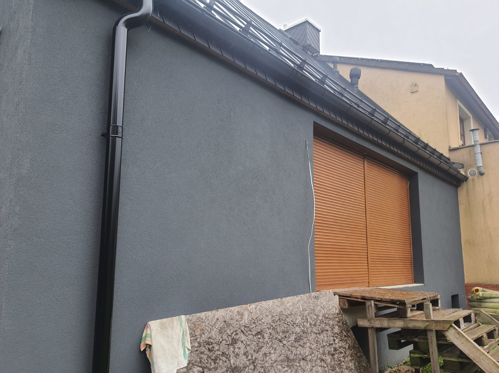 Świeżo otynkowana elewacja domu w kolorze grafitowym z zamontowanymi brązowymi roletami zewnętrznymi, widoczny fragment dachu z orynnowaniem i rura spustowa. Na pierwszym planie leży płyta OSB...