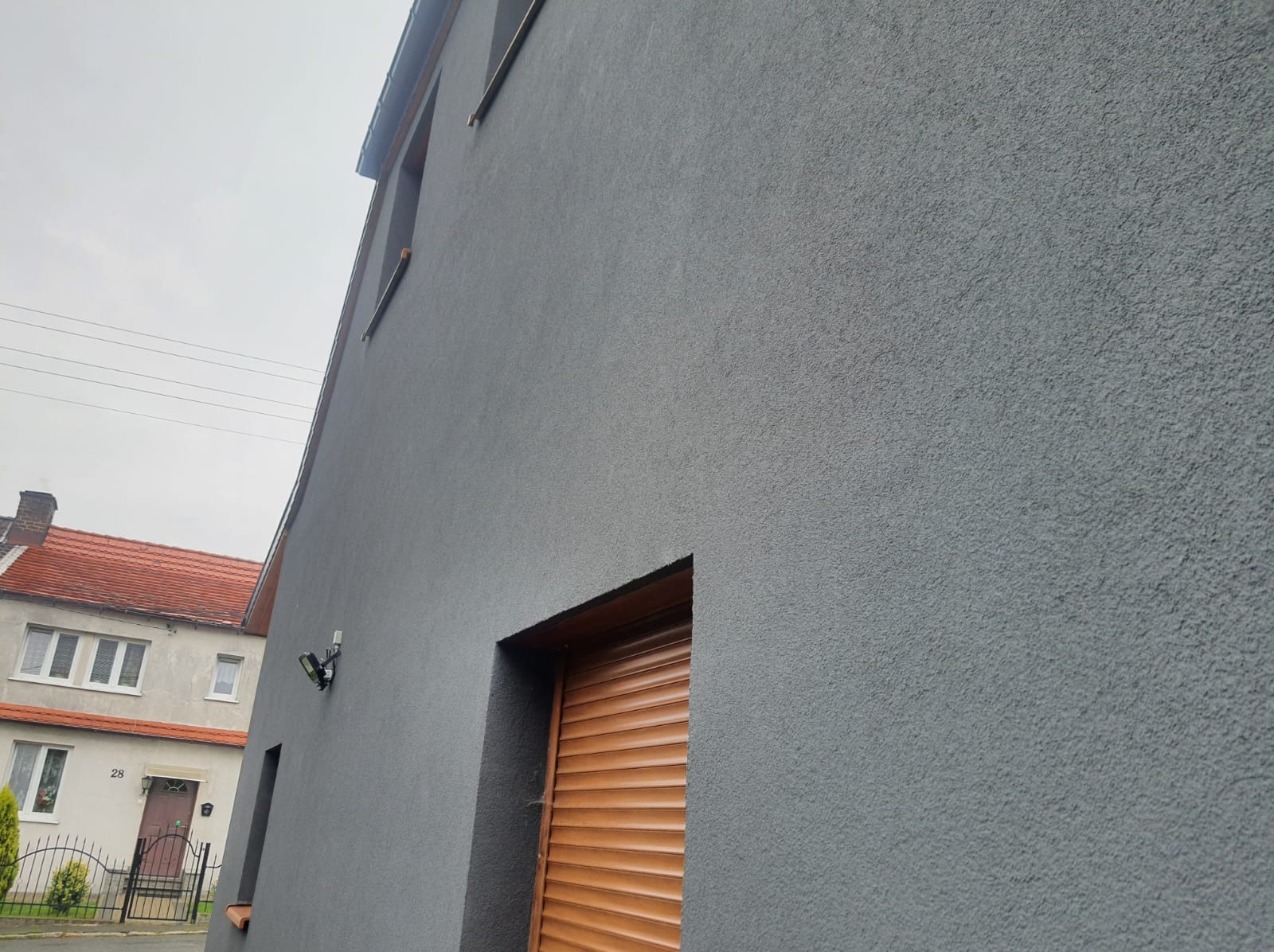 Szara elewacja budynku z fakturą baranka, widoczna brama garażowa z drewnianymi roletami, lampa zewnętrzna i fragment sąsiedniego domu z czerwoną dachówką.