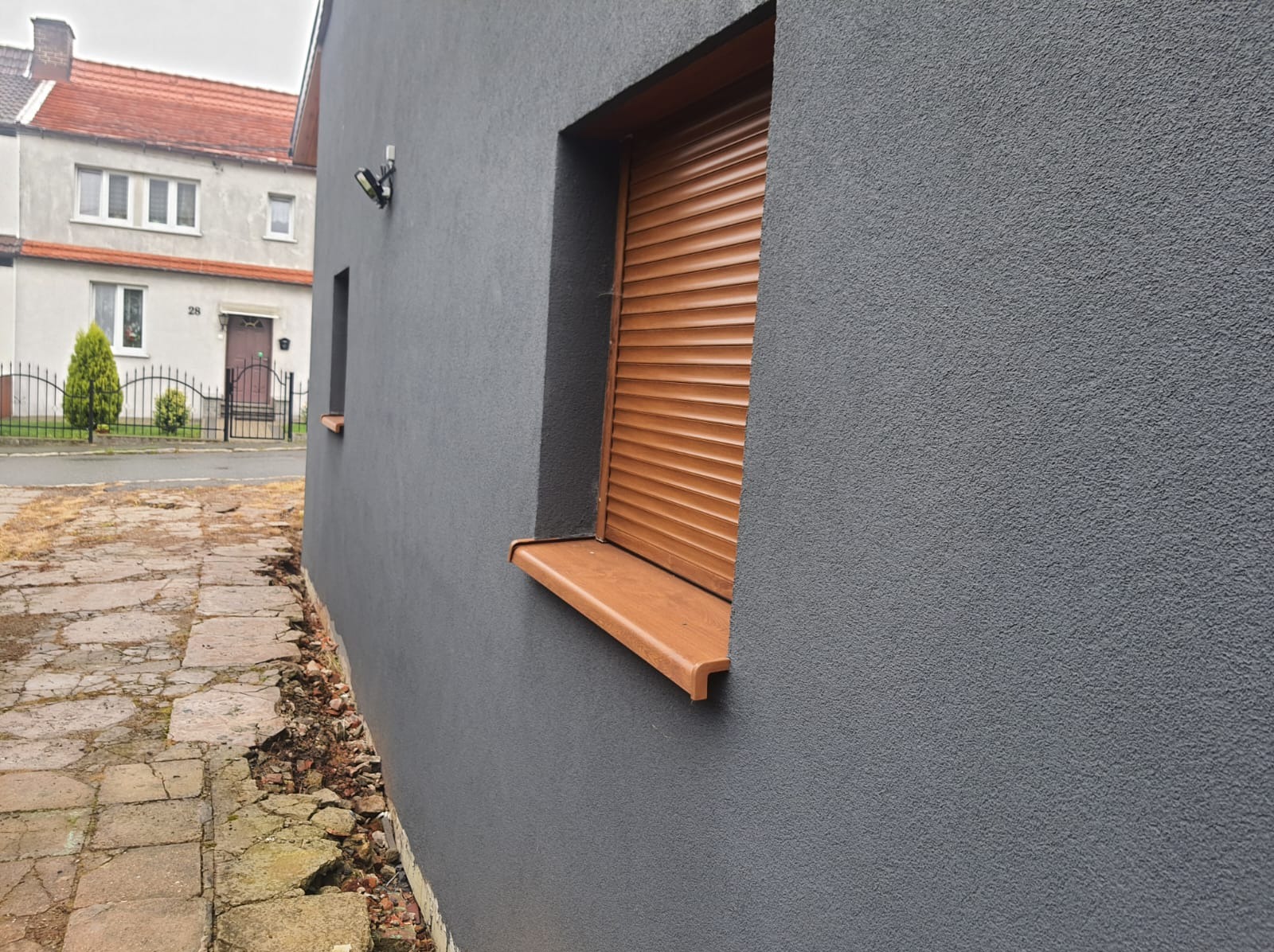 Elewacja budynku z szarą, ziarnistą strukturą, brązowe rolety zewnętrzne na oknach, widoczny fragment chodnika z popękanymi płytami.