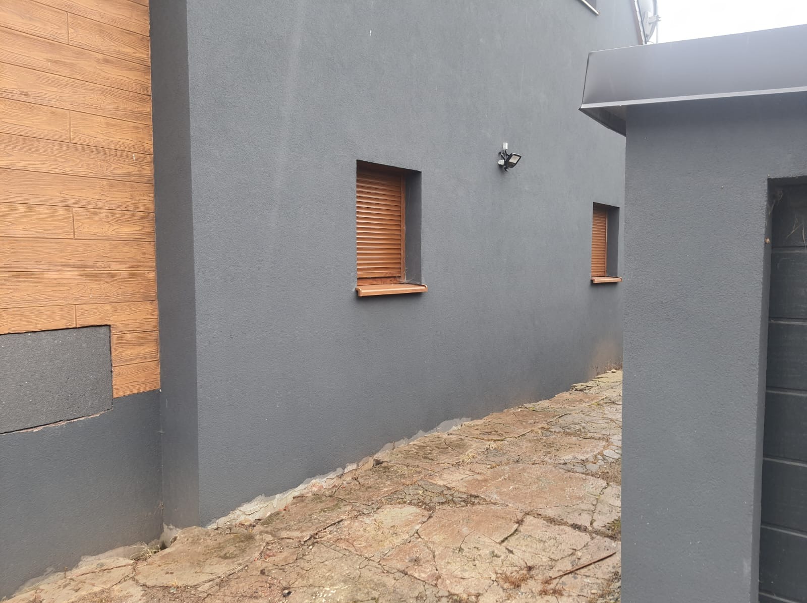 Szara elewacja budynku z dwoma oknami z brązowymi roletami zewnętrznymi, fragment drewnianej okładziny i kamienny chodnik.