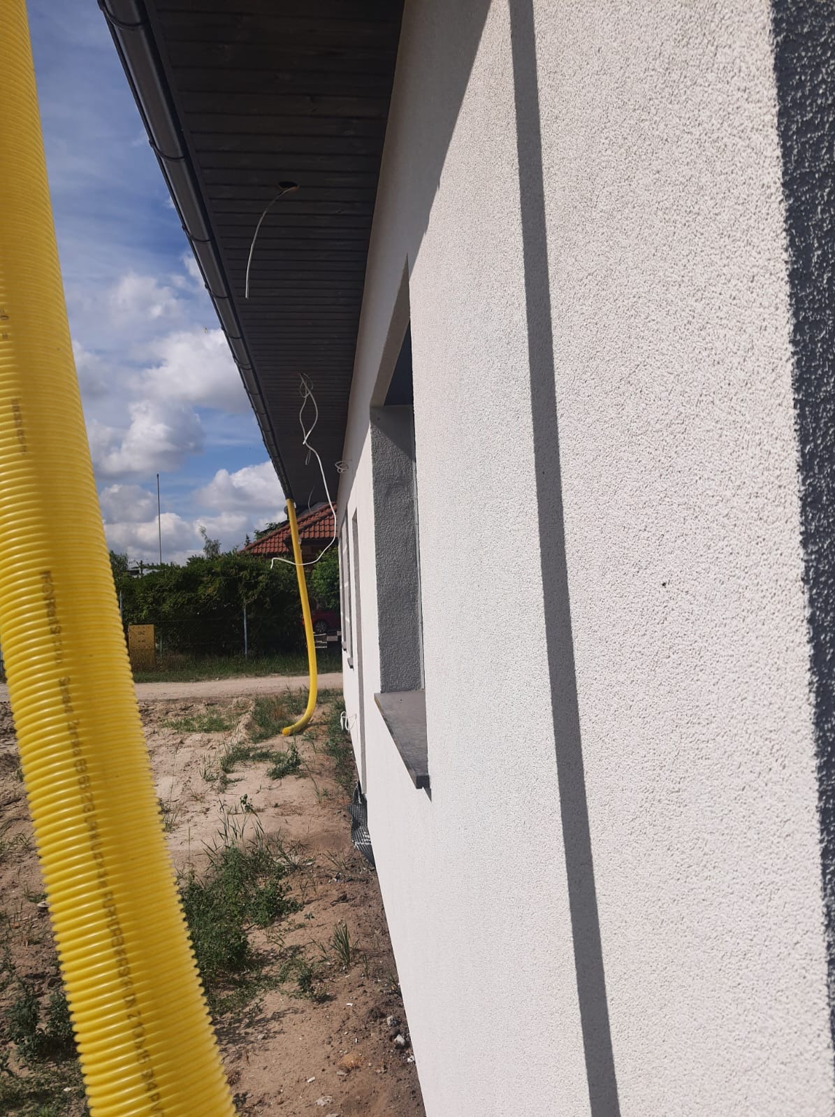 Fragment elewacji budynku z widoczną strukturą tynku, okno z szarą ramą, wystające przewody elektryczne spod dachu i żółta rura wentylacyjna na pierwszym planie.