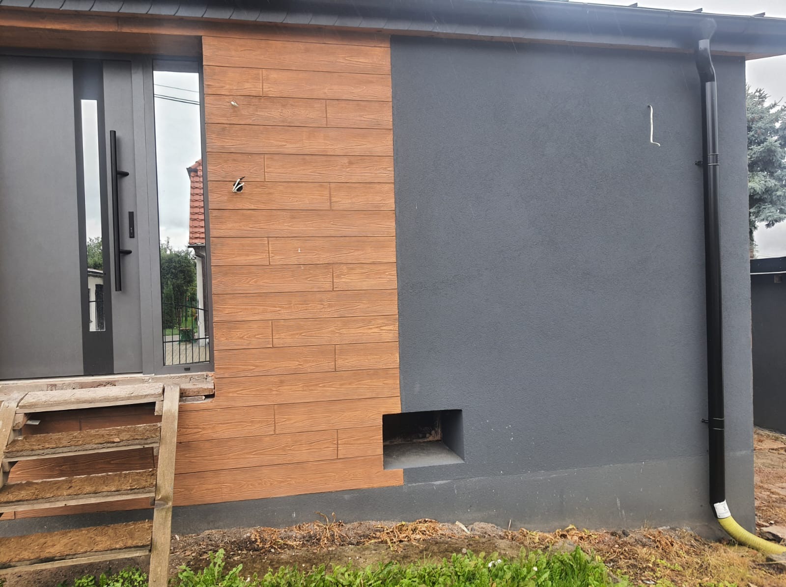 Fragment elewacji budynku z drewnopodobnymi panelami i szarym tynkiem, widoczne schody z drewna, rynna spustowa i niedokończone wykończenie wokół otworu wentylacyjnego.