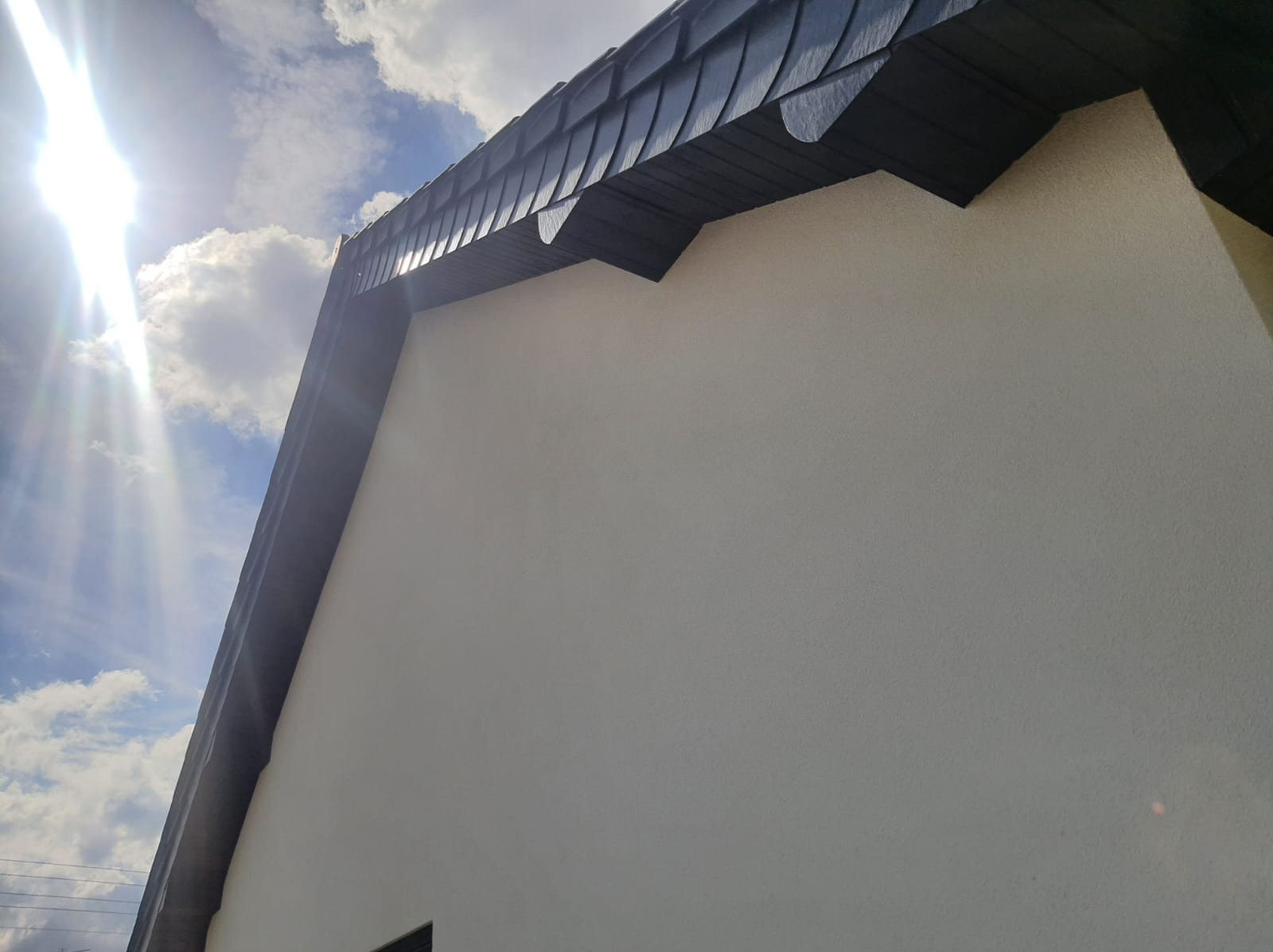 Fragment elewacji budynku z widocznym dachem pokrytym ciemną dachówką w kształcie łuski, oświetlonym jasnym słońcem i błękitnym niebem z chmurami w tle.