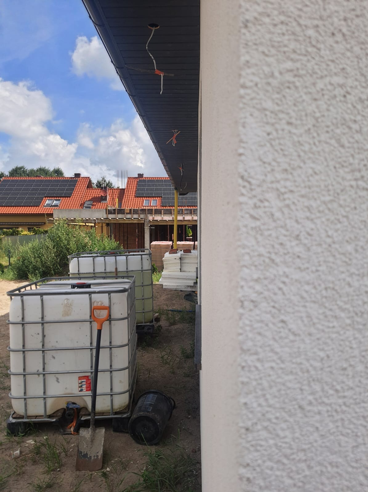 Ocieplanie elewacji budynku w trakcie prac: widoczne zbiorniki na wodę, szpachla, fragment otynkowanej ściany z fakturą baranka oraz wystające przewody pod dachem.