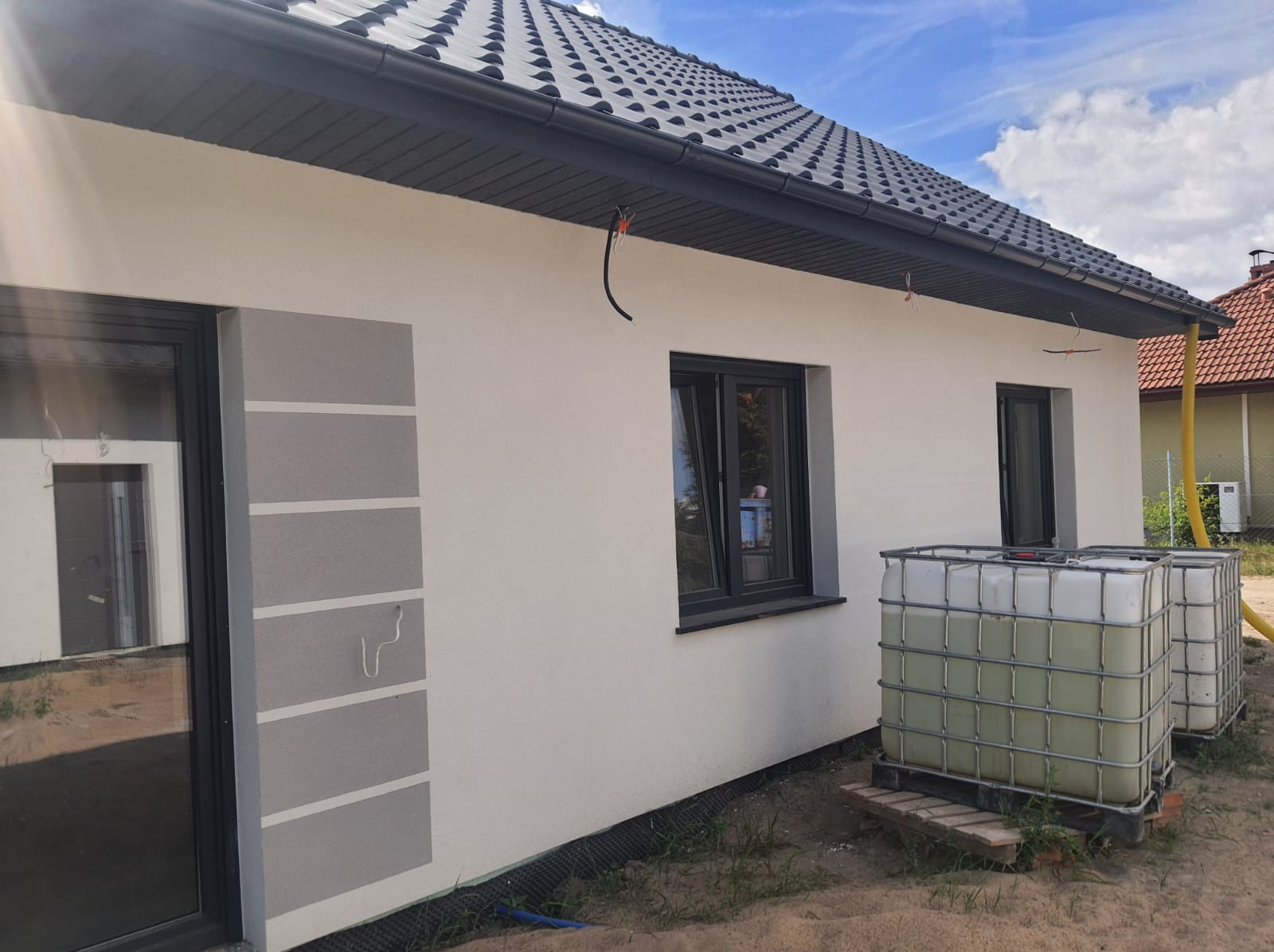 Widok z zewnątrz na dom jednorodzinny z szarą dachówką, białą elewacją z szarymi pasami dekoracyjnymi, czarnymi oknami i dwoma zbiornikami na wodę deszczową przy ścianie budynku.