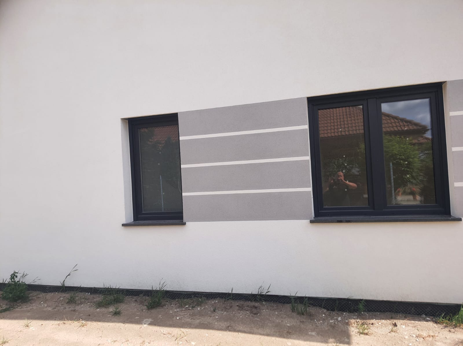 Elewacja budynku z dwoma czarnymi oknami, szarym pasem z białymi liniami i odbiciem fotografa w szybie.
