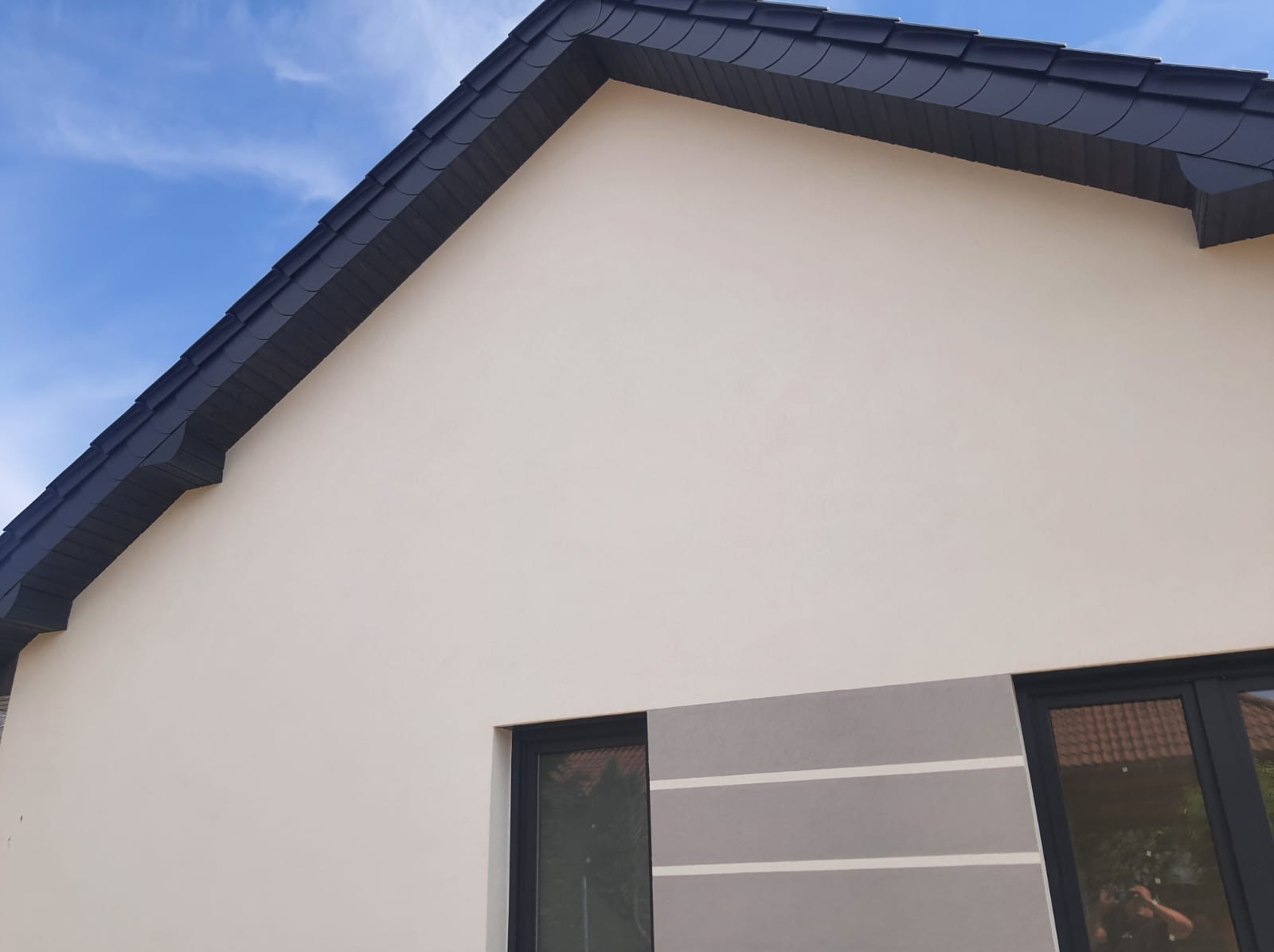 Fragment elewacji domu jednorodzinnego z widocznym dachem krytym ciemną dachówką, jasną elewacją i dekoracyjnym pasem szarości z białymi paskami pod oknem.