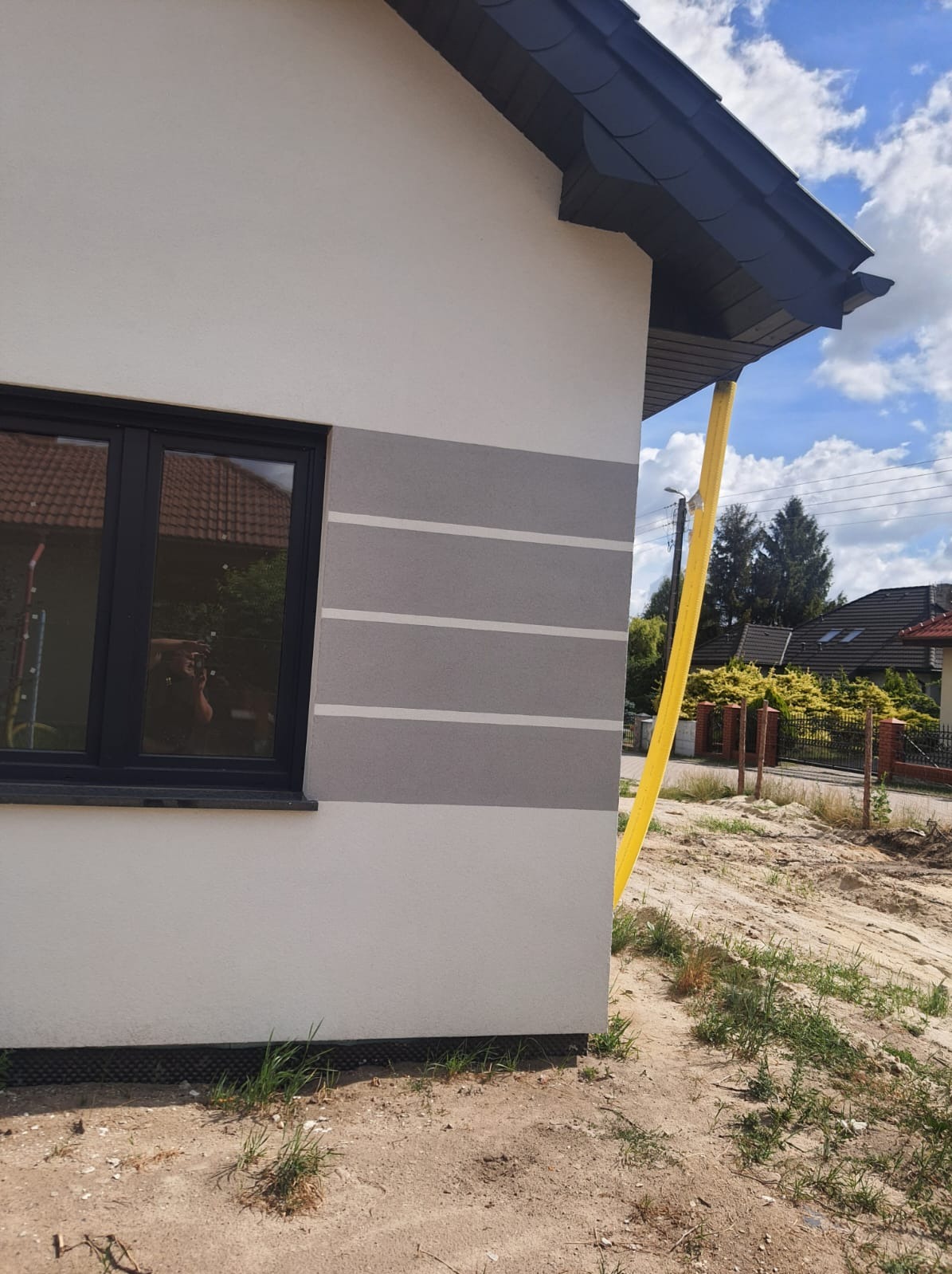 Fragment ściany domu po ociepleniu z poziomymi pasami w odcieniach szarości, widoczny narożnik dachu i okno z odbiciem fotografa, teren wokół domu w trakcie prac wykończeniowych.