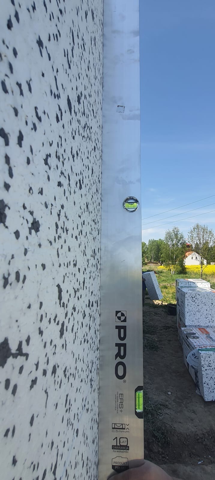 Ściana budynku pokryta styropianem, sprawdzana poziomicą marki PRO, z widocznymi paczkami styropianu w tle i fragmentem krajobrazu z domem.