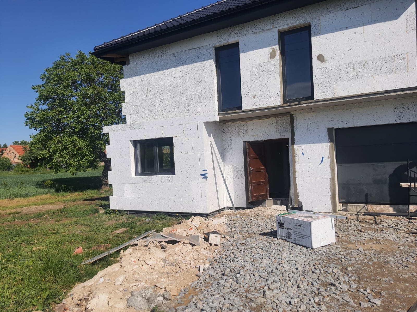 Surowy stan elewacji domu jednorodzinnego z białych bloczków, widoczne okna w ciemnych ramach, brama garażowa i częściowo wykonany dach, otoczenie placu budowy z gruzem i zielenią.