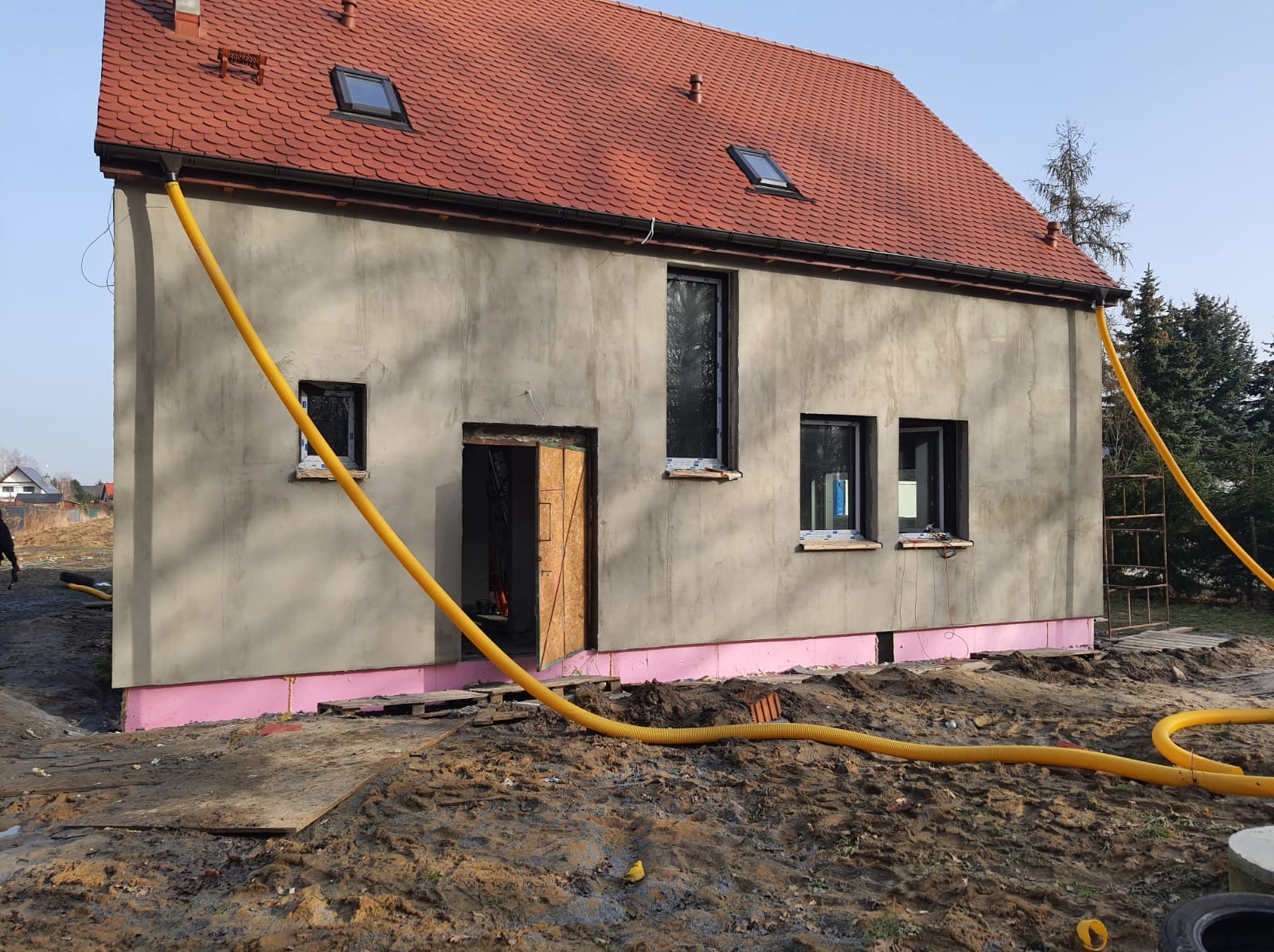 Dom jednorodzinny w trakcie ocieplania elewacji, widoczne styropianowe płyty fundamentowe, surowy tynk, okna z folią ochronną, dach z dachówką ceramiczną i żółty wąż doprowadzający materiał.