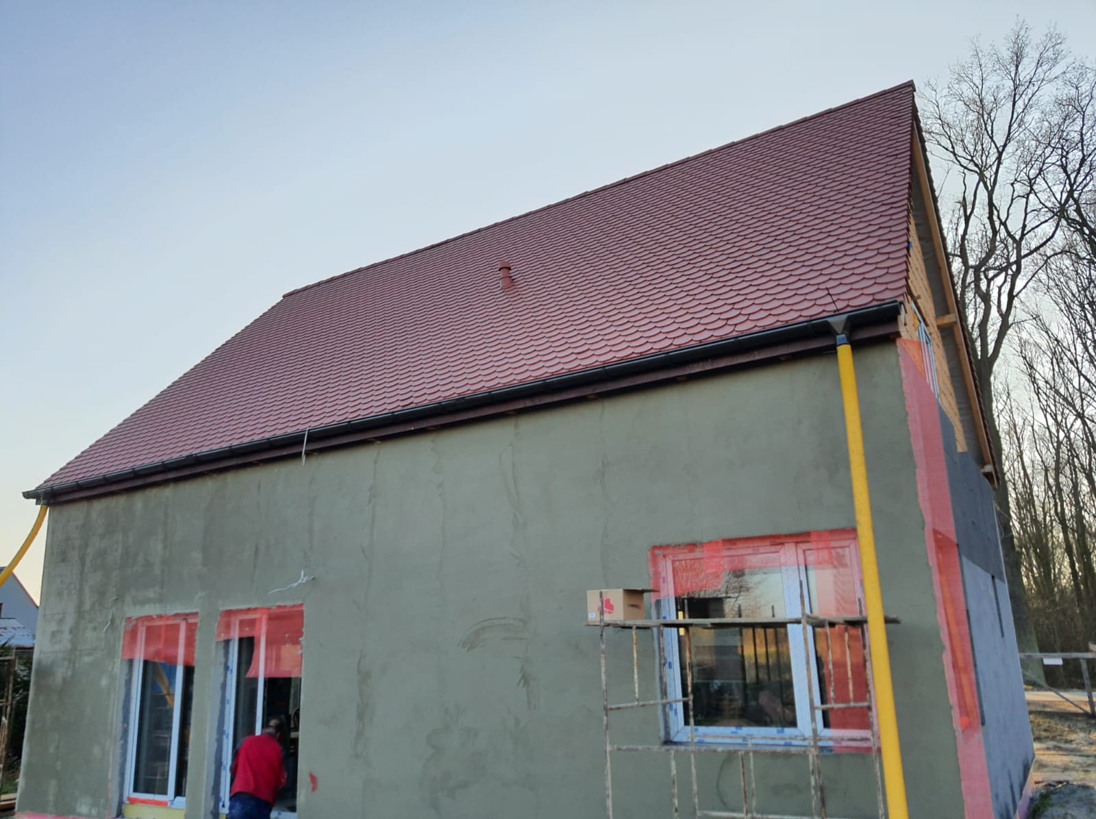 Budynek w Bydgoszczy z nowym, czerwonym dachem z dachówki, szarą elewacją w trakcie ocieplania, widoczne okna zabezpieczone folią i rusztowanie.