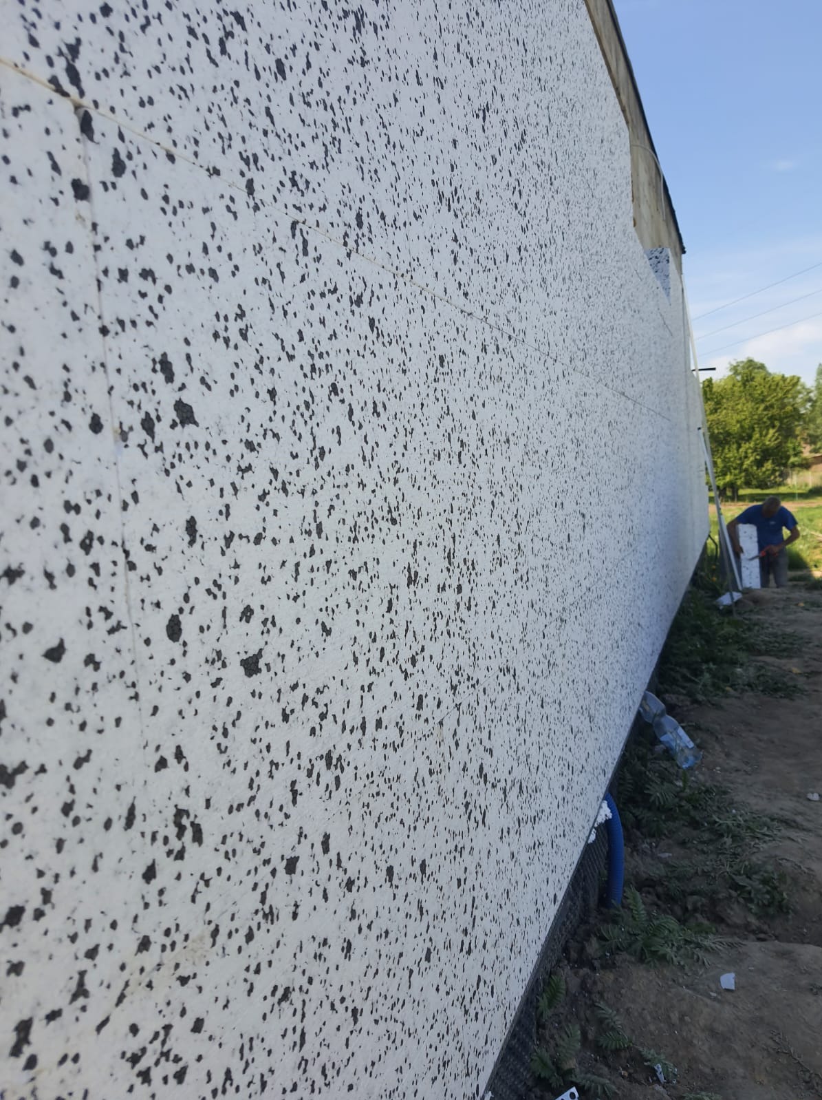 Ściana budynku w trakcie ocieplania, widoczne płyty styropianowe z nałożoną warstwą kleju z czarnymi plamkami, pracownik w oddali przycina płytę izolacyjną.