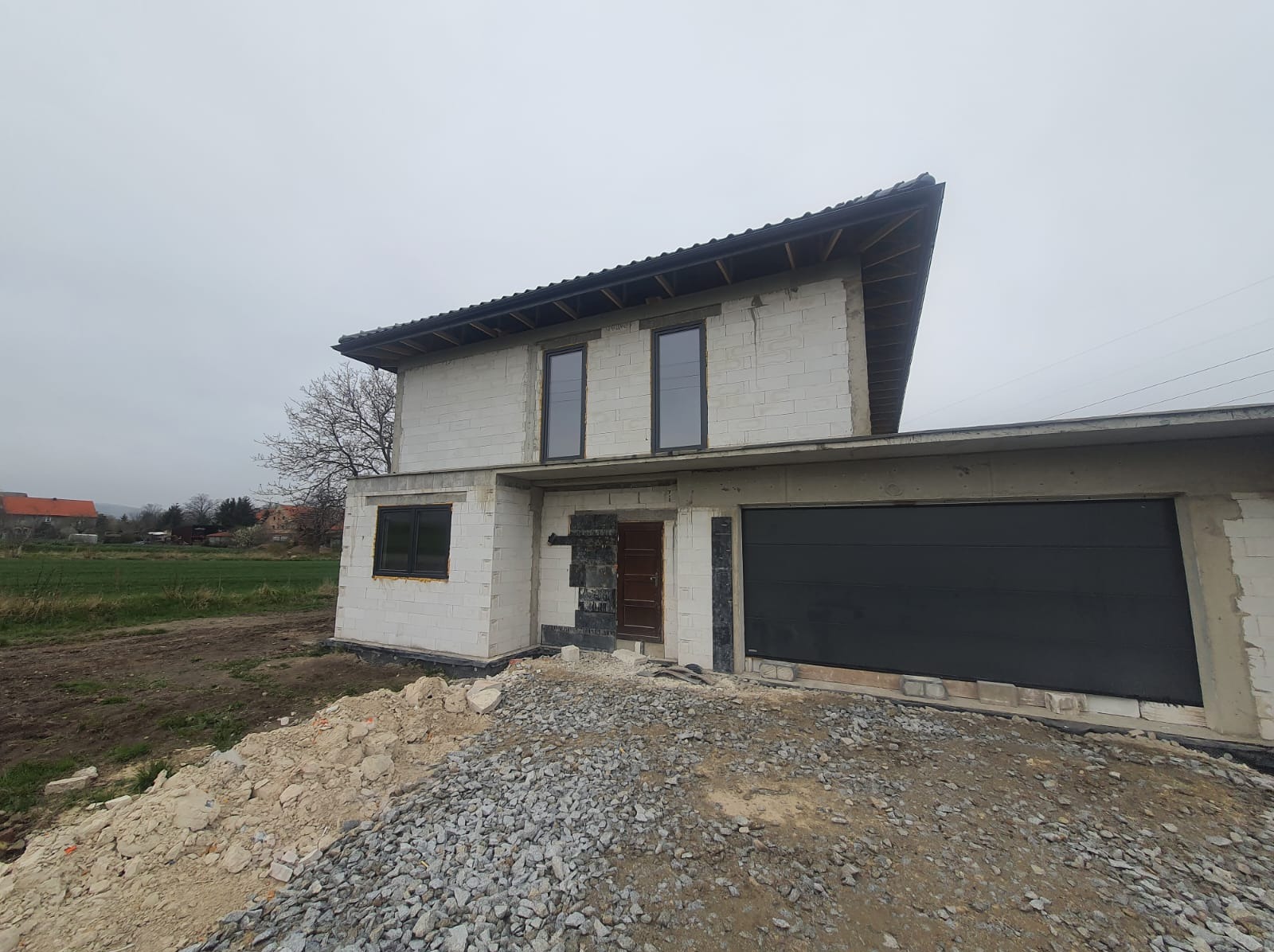 Surowy stan domu jednorodzinnego w trakcie budowy, widoczne białe bloczki, czarne okna i brama garażowa, na pierwszym planie gruz i ziemia, pochmurne niebo.