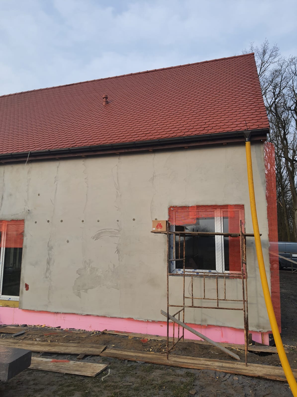 Dom w trakcie ocieplania elewacji z widocznym rusztowaniem, nowym dachem z dachówki ceramicznej i fragmentem żółtej rury wentylacyjnej.