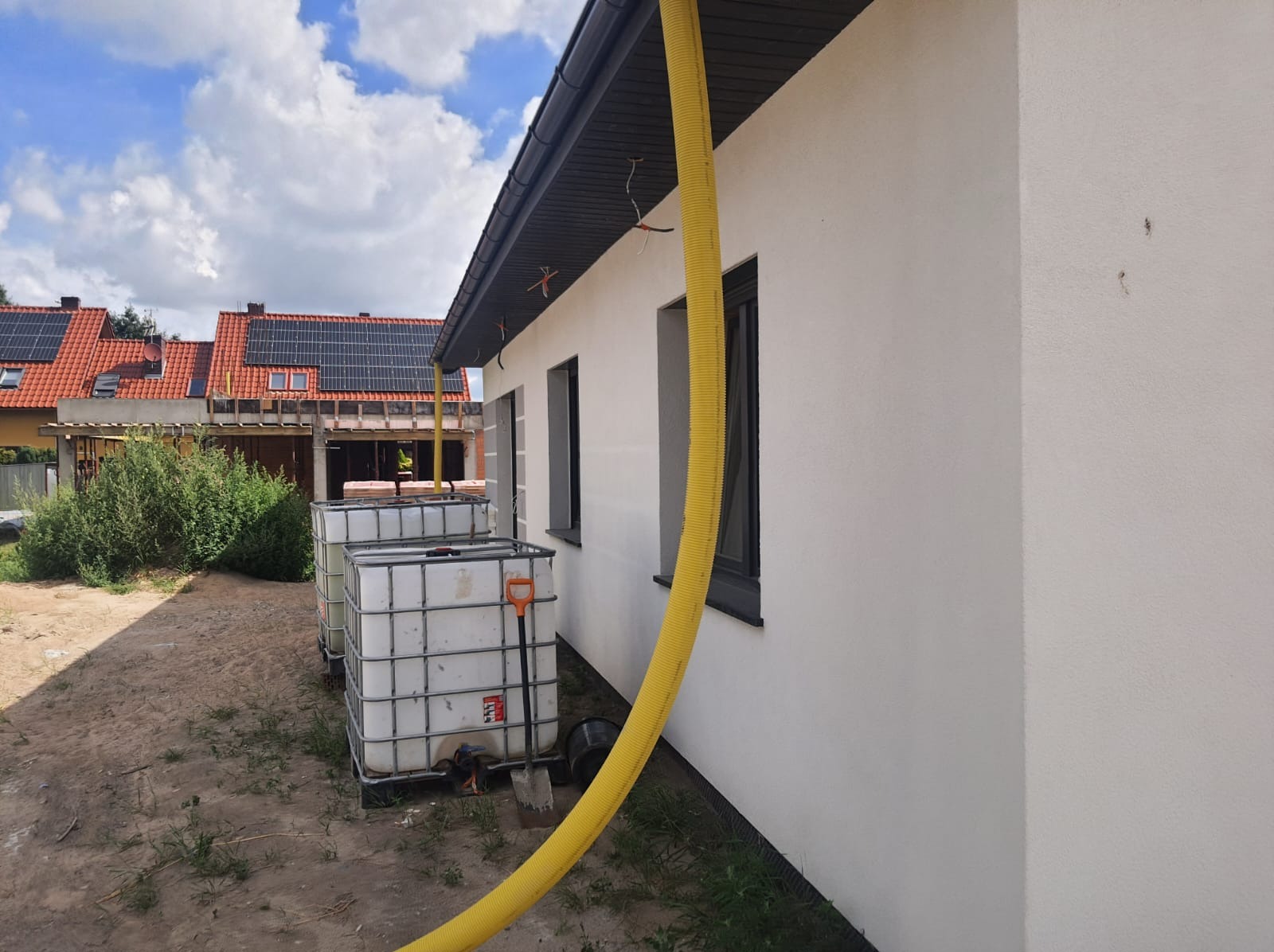 Świeżo otynkowany budynek z podłączonym żółtym wężem spustowym do zbiorników na deszczówkę, widoczne okna w szarych ramach, w tle budynek z panelami słonecznymi na dachu.