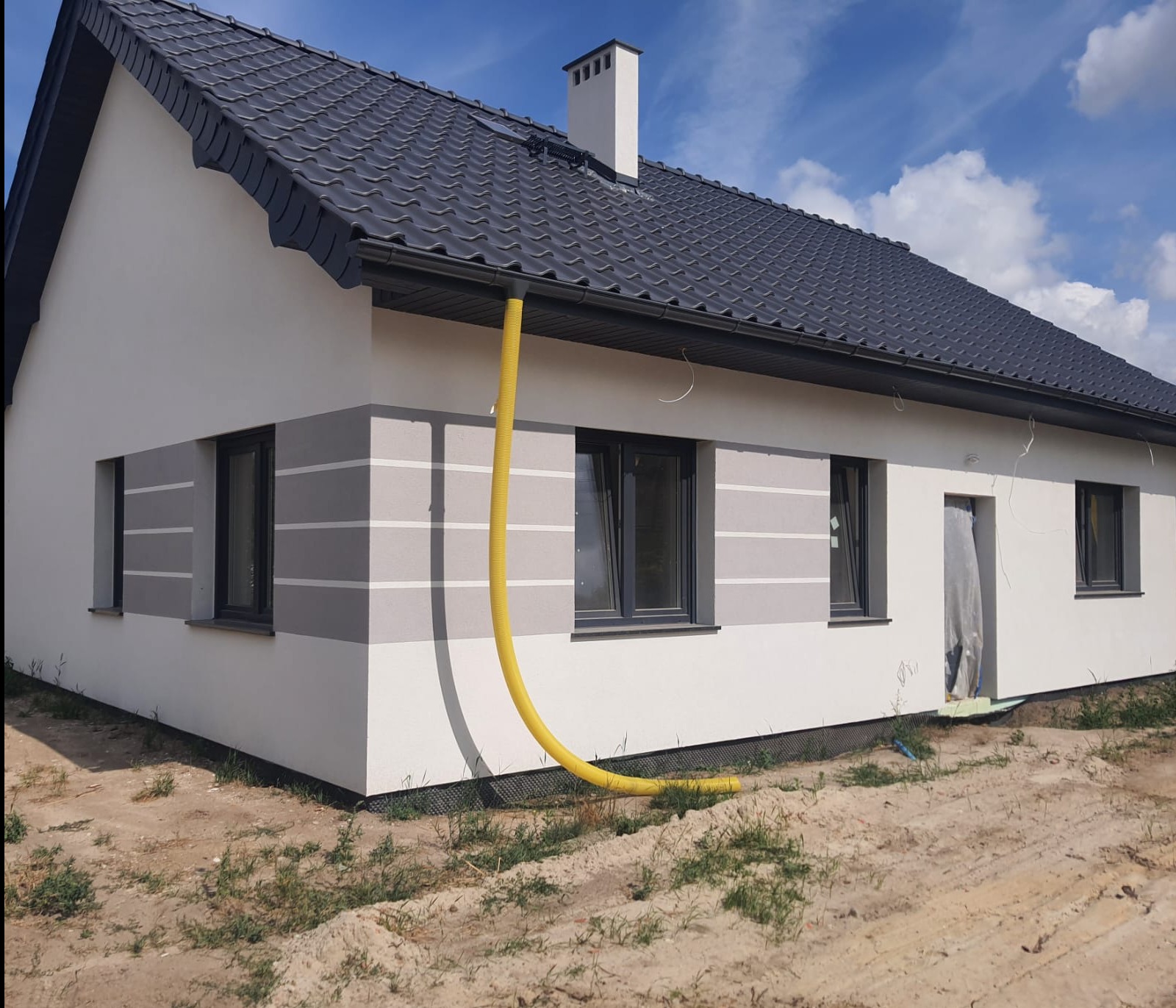 Nowo otynkowany dom jednorodzinny z ciemnoszarą dachówką, widoczny żółty wąż wentylacyjny przy elewacji, teren wokół domu w trakcie zagospodarowywania.