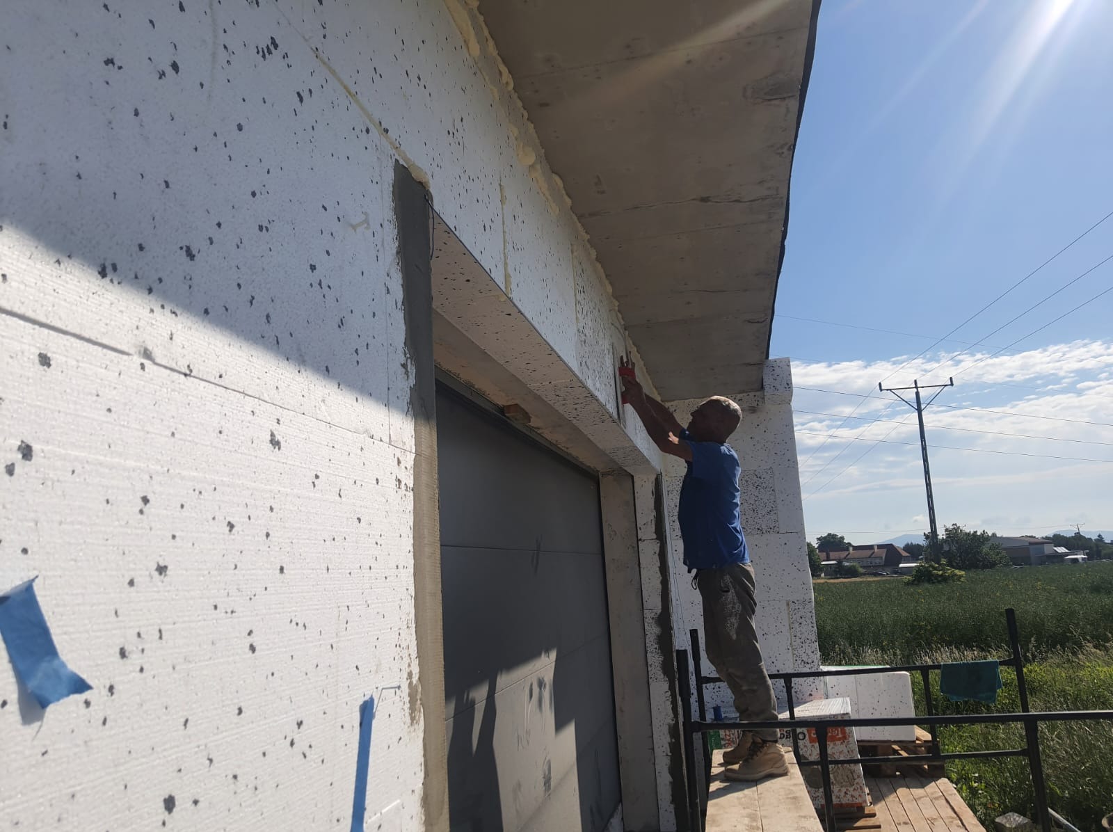 Pracownik na rusztowaniu wygładza piankę montażową wokół okna budynku ocieplonego styropianem, widoczne ślady po kołkach mocujących.