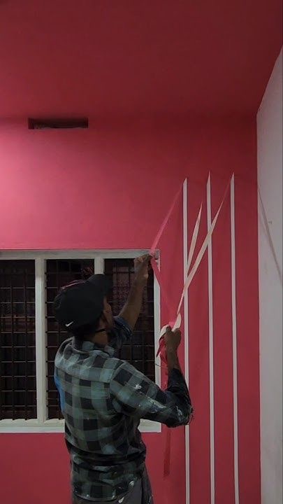 Malarz w trakcie wyklejania taśmą pionowych pasów na różowej ścianie, przygotowując się do malowania dekoracyjnego. Widoczne okno i fragment pomalowanej ściany.