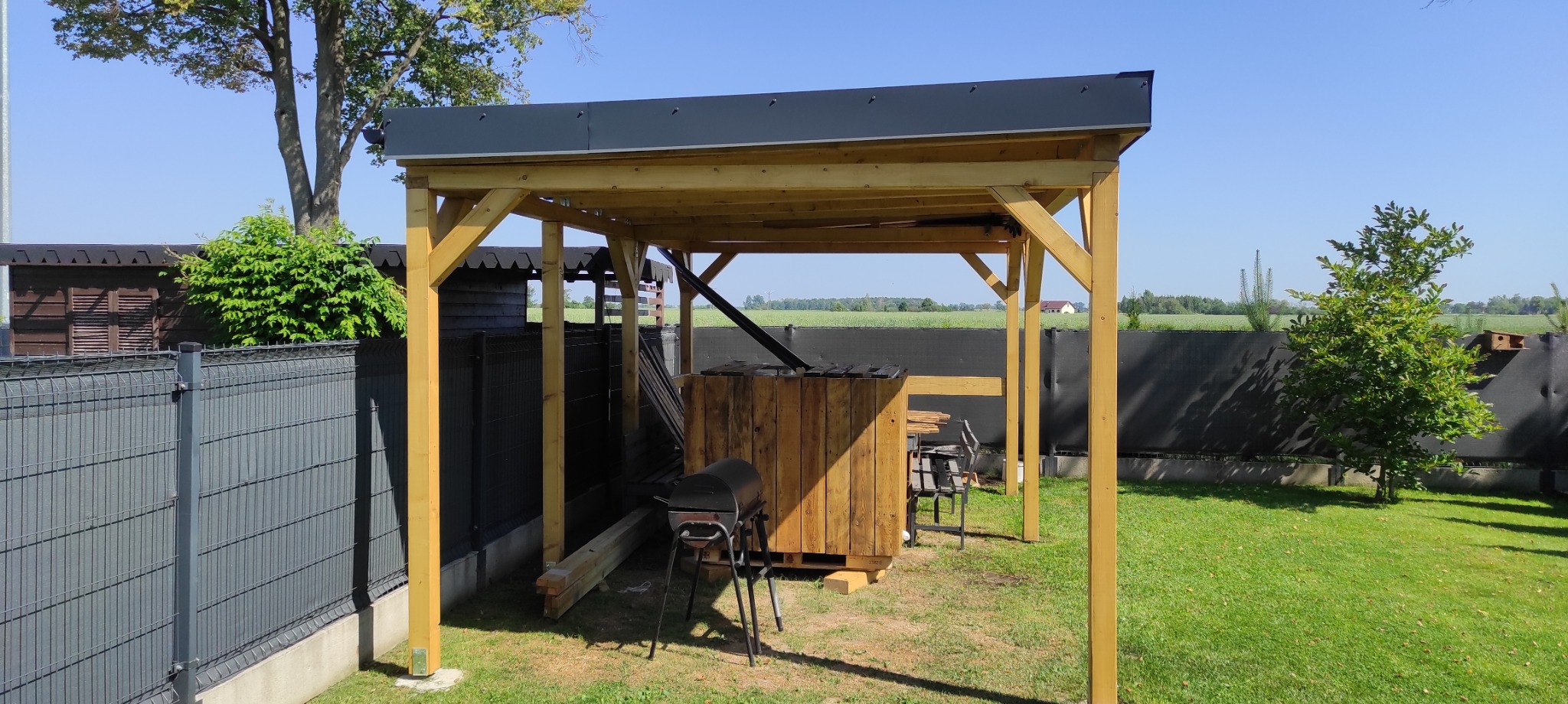 Surowa drewniana konstrukcja altany z płaskim, ciemnym dachem na tle zielonego trawnika i ogrodzenia, widoczne elementy grilla i drewniane skrzynie.