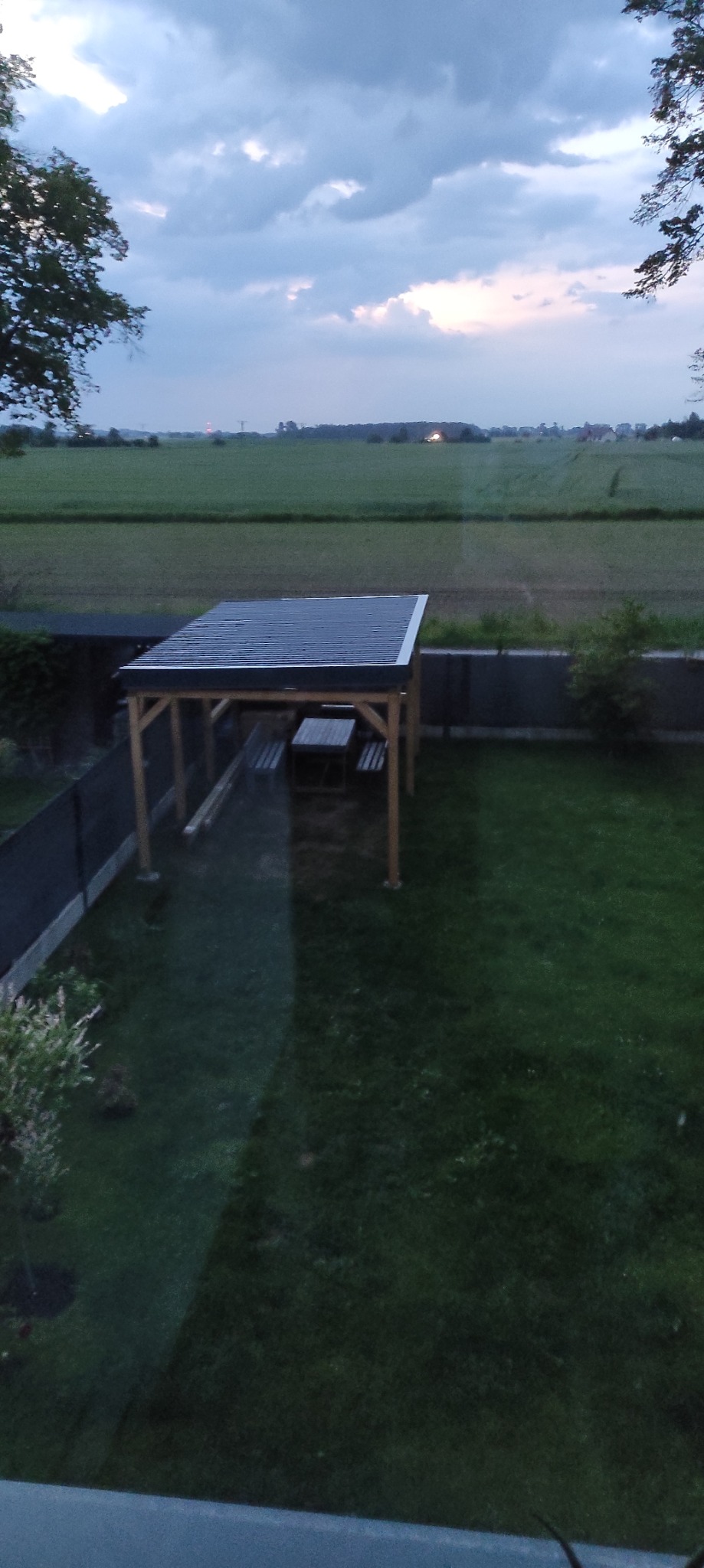 Zadaszona altana z drewnianą konstrukcją i stołem z ławkami, widok z góry na tle zielonego trawnika i pól uprawnych pod pochmurnym niebem o zmierzchu.