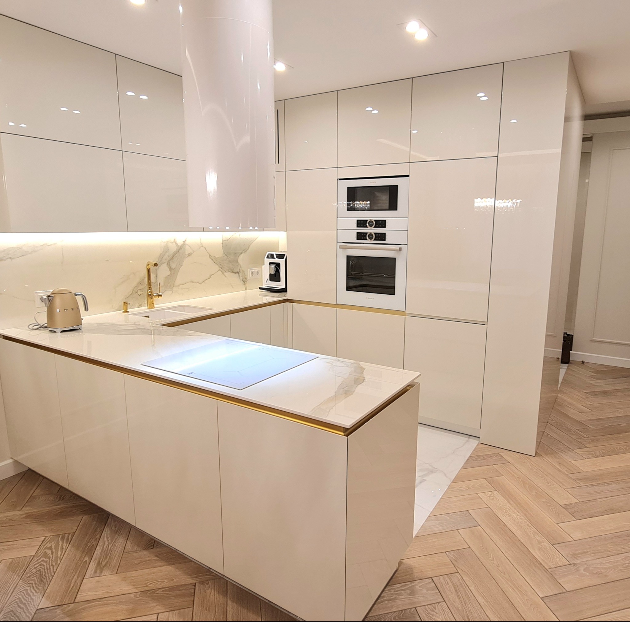 Nowoczesna kuchnia z białymi, lakierowanymi frontami, marmurowym blatem i złotymi akcentami, wyspą kuchenną z płytą indukcyjną i drewnianą podłogą w jodełkę.