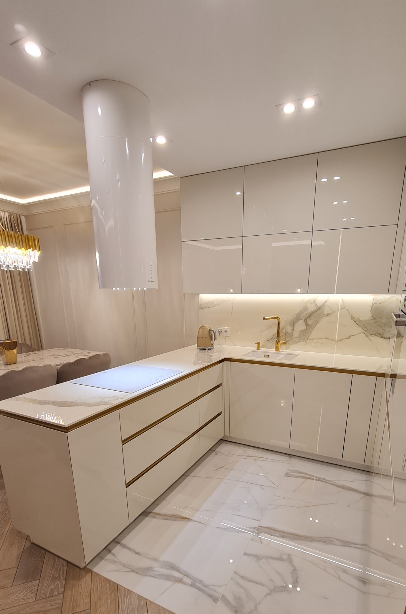 Nowoczesna, biała kuchnia na wymiar w Warszawie z połyskiem, z marmurowym blatem i podświetleniem LED, cylindryczny okap nad wyspą kuchenną, złote akcenty na szafkach i armaturze.