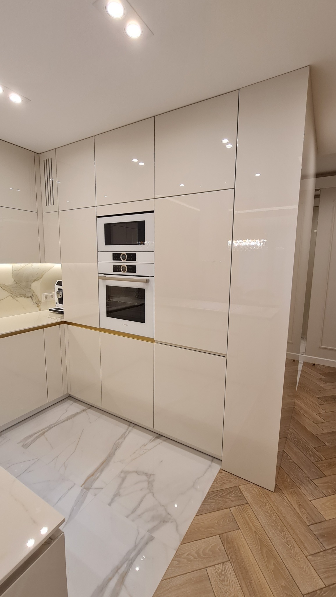 Nowoczesna kuchnia z białymi, lakierowanymi frontami, zabudowanym sprzętem AGD (piekarnik, mikrofalówka) i marmurową podłogą. Widoczny fragment parkietu ułożonego w jodełkę.