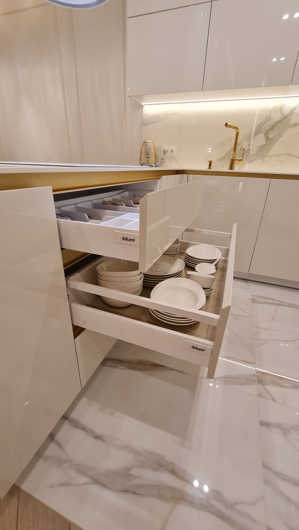 Biała kuchnia z marmurowym blatem, otwarte szuflady z systemem Blum wypełnione naczyniami, złota armatura i czajnik.