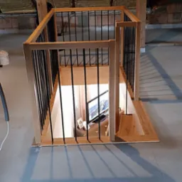 Otwór w stropie z drewnianą obudową i czarnymi, metalowymi balustradami, widok na pomieszczenie poniżej z telewizorem i meblami.