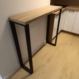 Blat z jasnego drewna na czarnych, metalowych nogach, przylegający do ściany w kuchni z białymi szafkami.