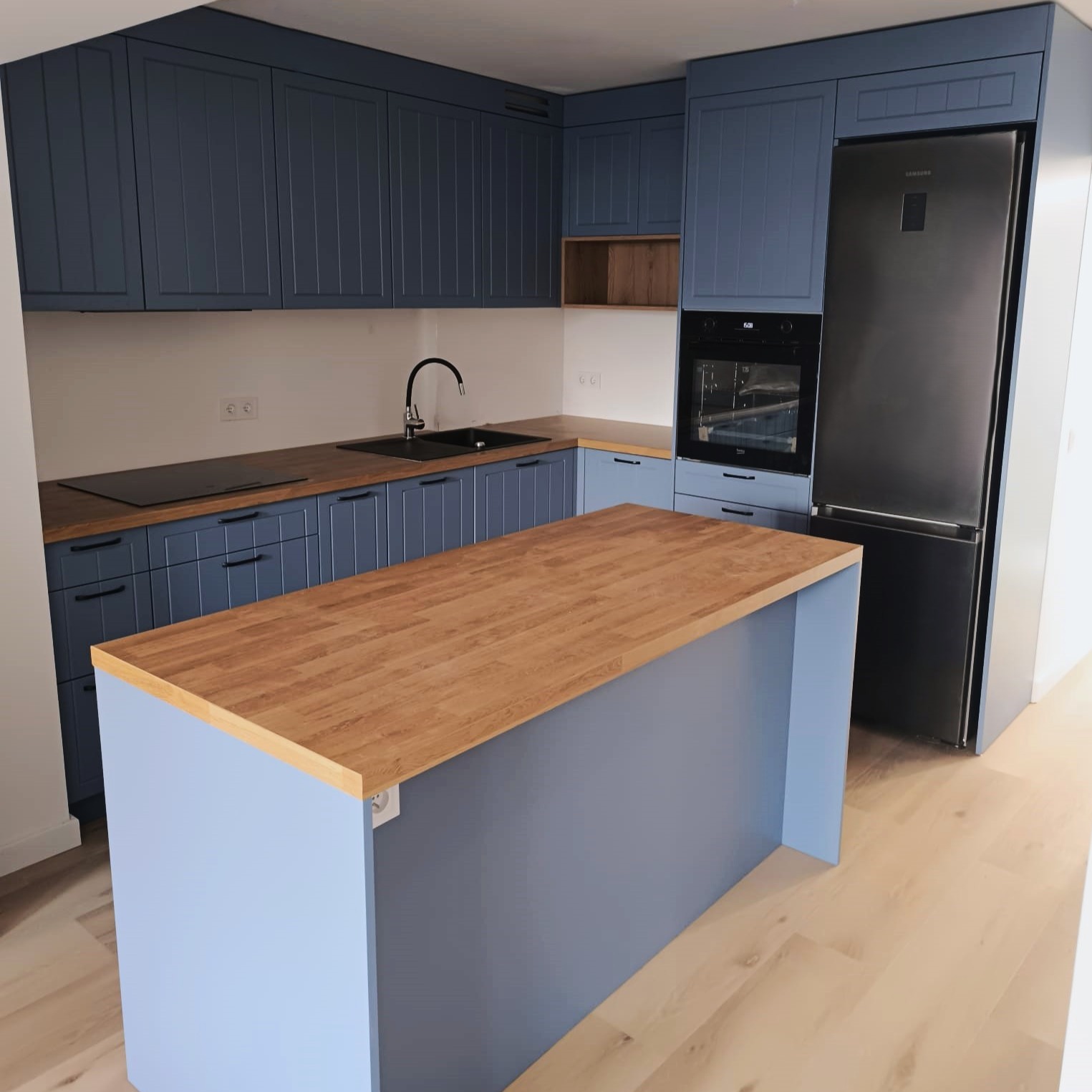 Kompletna kuchnia w odcieniach błękitu z drewnianym blatem wyspowym i sprzętem AGD w zabudowie, na jasnej drewnianej podłodze.