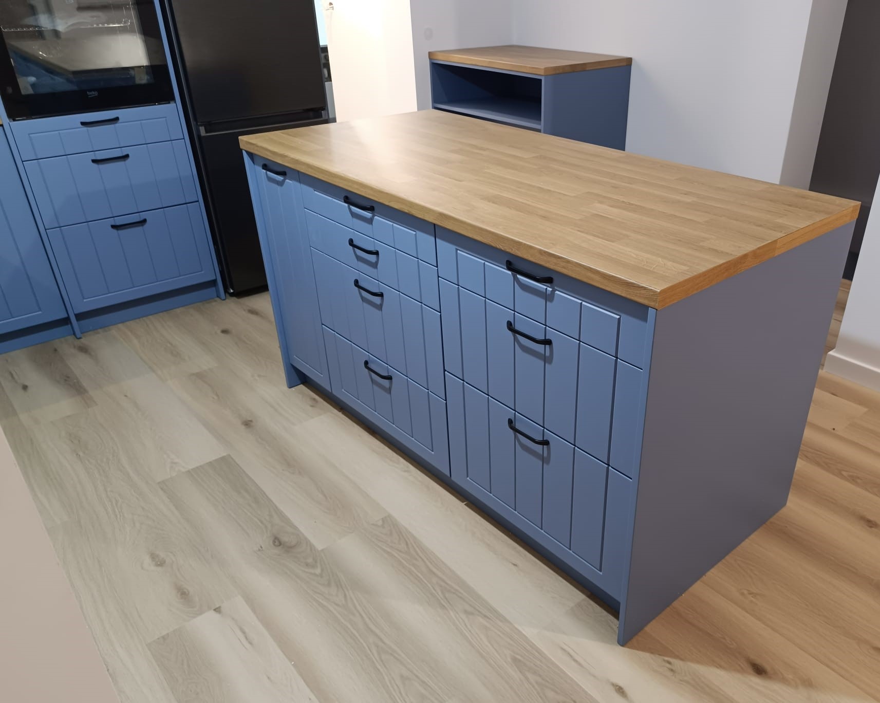 Niebieska wyspa kuchenna z drewnianym blatem i czarnymi uchwytami szuflad, uzupełniona o szafki kuchenne w tym samym kolorze i czarną lodówkę w tle.