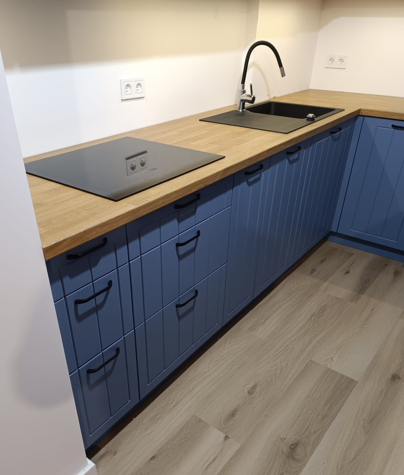 Fragment niebieskiej zabudowy kuchennej z drewnianym blatem, widoczny zlew, płyta indukcyjna i drewniana podłoga.
